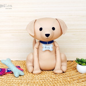 May include: A beige plush dog toy with black eyes and a blue collar. The collar has a bone-shaped tag that says "RUS". The dog is sitting on a woven rug with two bone-shaped toys in the foreground.