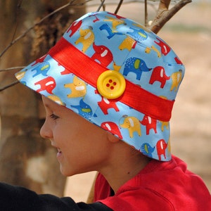 May include: A child wearing a blue and red sun hat with a yellow button and a pattern of colorful elephants. The hat has a red brim and a red band around the crown.