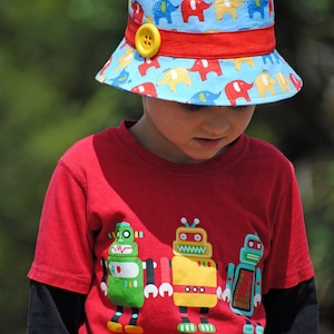 May include: A young child wearing a red t-shirt with a graphic of three robots. The child is wearing a blue and red bucket hat with a yellow button and a pattern of elephants.