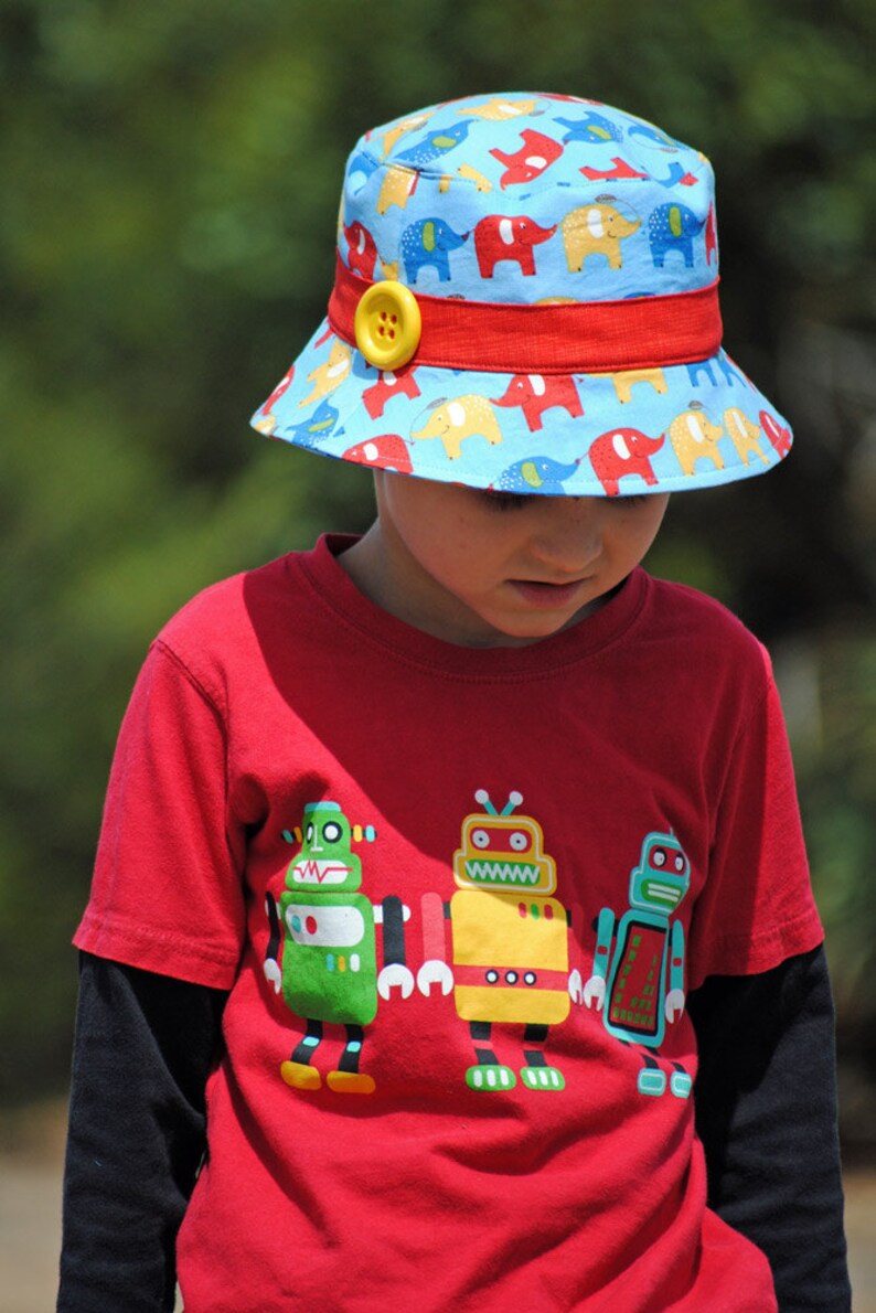 May include: A young child wearing a red t-shirt with a graphic of three robots. The child is wearing a blue and red bucket hat with a yellow button and a pattern of elephants.