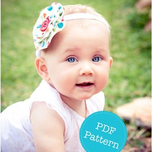 May include: A baby girl wearing a white dress with a headband featuring a colorful fabric flower. The headband is white with a blue polka dot pattern. The baby is looking at the camera and smiling. The image has a blue circle with the text "PDF Pattern" in white.