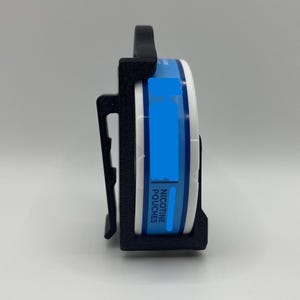 May include: A black 3D-printed holder for a round container of nicotine pouches. The container is white with a blue label and the words "NICOTINE POUCHES". The holder has a clip for attaching to a belt or pocket.