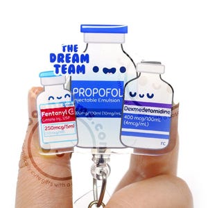 May include: Three cartoon vials of medication: Fentanyl, Propofol, and Dexmedetomidine.  The vials are attached to a retractable badge reel. The text 'The Dream Team' is featured above the Propofol vial.  The vials are predominantly white with blue caps and labels containing medication information and dosages. The design is playful and whimsical.