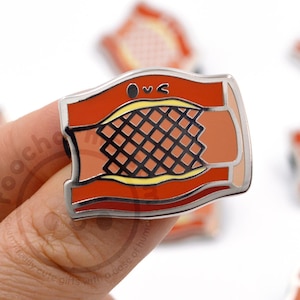 May include: A rectangular enamel pin featuring a stylized bacon design with a silver border. The pin has a red and orange color scheme with a black grid pattern in the center. The pin is held by a person's fingers.