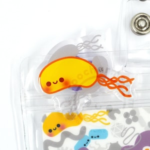May include: A clear plastic badge holder with a yellow cartoon bacteria with a smiling face and orange flagella. The badge holder has a clear plastic strap with a silver button.