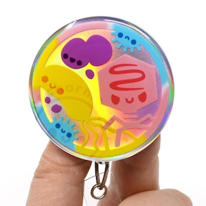 May include: A round, iridescent badge with a cartoon design featuring various colorful shapes and characters. The badge has a clear plastic covering and a metal clip on the back. The design includes the word "learn".