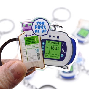 May include: A clear acrylic keychain with a cartoon illustration of a feeding tube and a medical pump. The feeding tube has a label that reads "Complete Balanced Nutrition with Fiber" and "1.5 Cal". The medical pump has a screen that reads "Running" and "Feed Rate 150 ml/hr". The keychain also has the text "You Fuel Me Up".