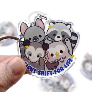 May include: A clear acrylic keychain with a group of cartoon animals. The animals are a raccoon, an owl, a bat, and a possum. The keychain has the text "Night Shift for Life" in blue letters.