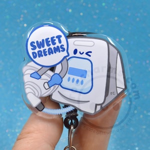 May include: A clear, cartoon-style badge reel featuring a white and gray medical device with a smiling face. A speech bubble says "Sweet Dreams" in blue. The reel has a black bead chain and a silver clip.