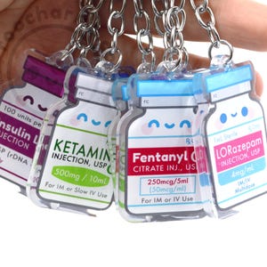 May include: A collection of acrylic keychains shaped like medicine bottles, each with a different label and a smiling face. The labels include "Insulin L", "Ketamine", "Fentanyl", and "Lorazepam". Each keychain has a silver chain.
