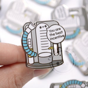 May include: A silver enamel pin featuring a cartoon illustration of a medical breathing machine with a blue tube and a speech bubble that says "You are the best incentive".