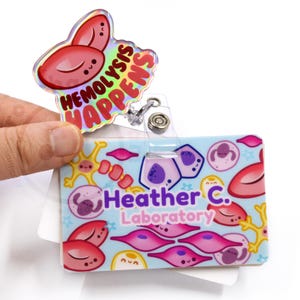May include: A blue and white ID badge with a cartoon illustration of cells and the text "Heather C. Laboratory". A holographic badge holder with the text "Hemolysis Happens" is attached to the badge.