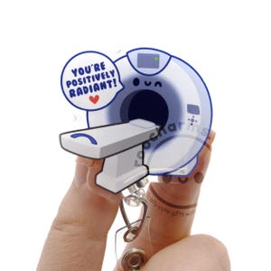 May include: A clear acrylic badge reel featuring a cartoon illustration of a white and blue CT scanner with a patient table. A blue speech bubble above the scanner contains the text YOU'RE POSITIVELY RADIANT! with a small red heart icon.