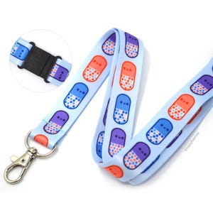 May include: A light blue lanyard featuring a repeating pattern of smiling pill capsules in shades of orange, blue, and purple. The lanyard includes a silver metal lobster clasp and a black plastic breakaway buckle, with the text roocharms printed on it.