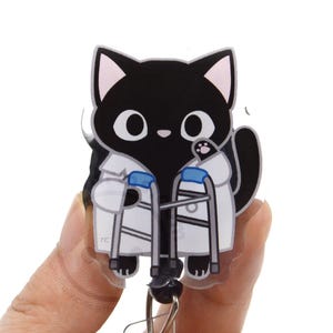 May include: An acrylic badge reel featuring a black cat with large white eyes wearing a white lab coat and holding a grey medical walker with blue accents. The cat has pink inner ears and a small pink nose, with the letters rc printed on the bottom left.