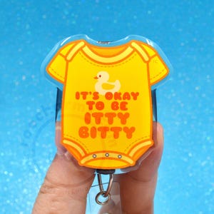 May include: A yellow badge reel shaped like a baby onesie featuring a small white duck graphic and the orange text IT'S OKAY TO BE ITTY BITTY. The item has a clear plastic casing and a metal clip attachment at the bottom for securing to clothing.