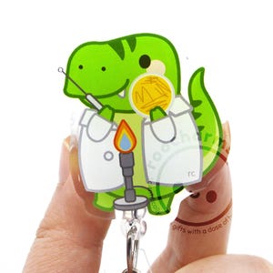 May include: A green T-Rex character wearing a white lab coat, holding a yellow petri dish and a metal inoculating loop. In front of the dinosaur is a lit Bunsen burner with a blue and orange flame. The design is printed on a clear acrylic badge reel.