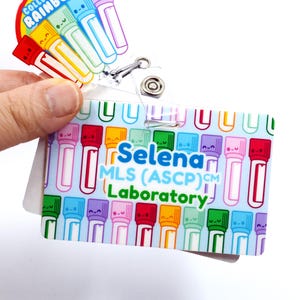 May include: A blue and white ID badge with a colorful pattern of cartoon test tubes. The text "Selena MLS (ASCP)CM Laboratory" is printed on the badge.