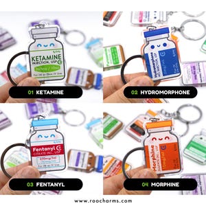 May include: A collection of four acrylic keychains shaped like medicine bottles, each with a different label: Ketamine, Hydromorphone, Fentanyl, and Morphine. The keychains have black key rings and are held against a blurred background.