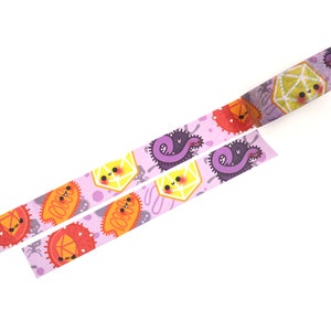 Op de afbeelding: Roze washi tape met een herhalend patroon van schattige cartoon-microben, waaronder een paarse worm, een gele zeshoek en een oranje ovaal. De tape is uitgerold en toont het herhalende patroon.