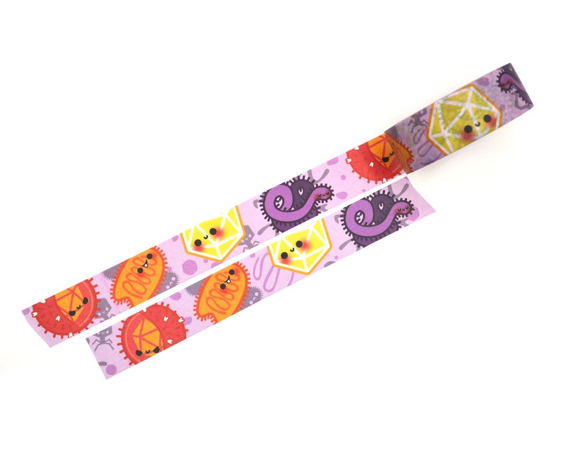 Op de afbeelding: Roze washi tape met een herhalend patroon van schattige cartoon-microben, waaronder een paarse worm, een gele zeshoek en een oranje ovaal. De tape is uitgerold en toont het herhalende patroon.