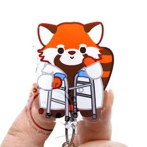 May include: An acrylic badge reel featuring a cartoon red panda wearing a white lab coat and holding a silver medical walker. The red panda has orange and white fur, brown markings, and a striped tail. A circular logo is visible on the lower left.
