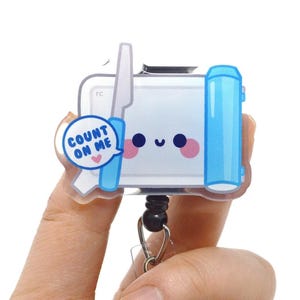 May include: A badge reel featuring a rectangular white character with a smiling face, pink cheeks, and a blue knife. A speech bubble contains the text 'COUNT ON ME' with a small pink heart. The design is printed on a clear acrylic base.
