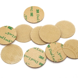 May include: A collection of ten circular 3M Dual Lock fasteners. Each piece features a tan, textured hook-and-loop surface on one side and a tan adhesive backing on the other, with the green text 3M Dual Lock printed on the adhesive side.