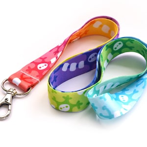 May include: A rainbow lanyard with a silver clip. The lanyard is made of a soft, colorful fabric with a repeating pattern of white skeletons.