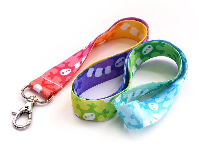 May include: A rainbow lanyard with a silver clip. The lanyard is made of a soft, colorful fabric with a repeating pattern of white skeletons.