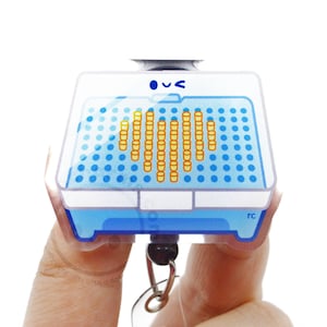 May include: A clear acrylic badge reel featuring a blue rectangular tray graphic with a grid of blue dots and a central heart shape formed by orange dots. The top of the tray has a small blue winking face icon and the letters rc in the corner.