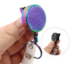 May include: A retractable badge reel featuring a circular housing with an iridescent rainbow finish and a textured purple glitter surface. The device includes a metal clip, a split ring, and a clear plastic strap with two metal snaps for attachment.
