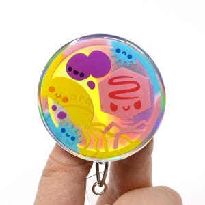 May include: A circular badge reel featuring a holographic background with colorful cartoon illustrations of various microbes and a bacteriophage. The design includes a yellow, blue, and purple color palette with smiling faces on each character.