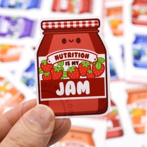 May include: A red and white cartoon jar of jam with a smiling face. The jar has a white label with the text "Nutrition is my jam" and three red strawberries on the label.