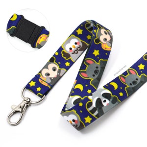 May include: A blue lanyard with a white background and a repeating pattern of cartoon animals, including an owl, a raccoon, a bat, and a mouse. The lanyard has a silver clip and a black adjustable buckle. The text "@roocharms" is printed on the lanyard.