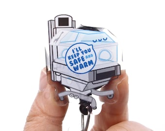 May include: A clear acrylic badge reel featuring a graphic illustration of a neonatal incubator. The design includes a blue speech bubble with the text I'LL KEEP YOU SAFE AND WARM, a small giraffe icon, and a metallic clip mechanism at the base.