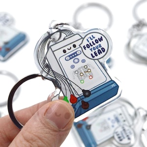 May include: A clear acrylic keychain featuring a cartoon illustration of a medical heart monitor with a smiling face. The keychain has a silver ring and a small round charm that says "I'll follow your lead".