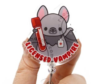 Puede incluir: Un portatarjetas circular de acrílico con un murciélago gris de dibujos animados con orejas rosadas y pequeños colmillos. El murciélago sostiene un tubo de ensayo con tapa roja lleno de líquido rojo y lleva una etiqueta médica blanca con una cruz roja.
