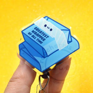 May include: A blue acrylic badge reel featuring a cartoon illustration of a candy wrapper with a smiling face. The text GREATEST WRAPPER OF ALL TIME is printed in dark blue on the front of the light blue rectangular wrapper design.