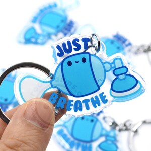 May include: A clear blue and white acrylic keychain with a cartoon illustration of a blue whale wearing an oxygen mask and the text "Just Breathe".