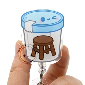 May include: A clear plastic badge reel featuring a cartoon illustration of a brown wooden stool inside a specimen container with a light blue lid. The lid has a winking face with pink cheeks. The text roocharms.com is printed on the container.
