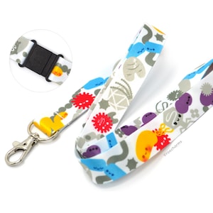 May include: A white fabric lanyard featuring a colorful pattern of cartoon microbes, bacteria, and viruses in shades of red, blue, yellow, and purple. The lanyard includes a silver metal lobster clasp and a black plastic breakaway buckle.