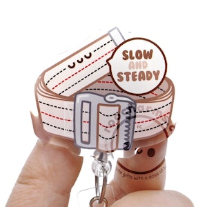 May include: A badge reel featuring a beige strap with red and black dashed lines, a grey buckle, and a speech bubble with the text SLOW AND STEADY. The design is printed on a clear acrylic piece attached to a retractable cord and a clear plastic clip.