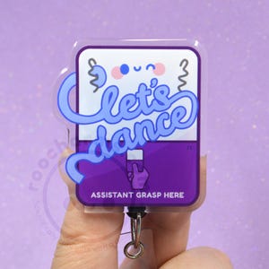 May include: A rectangular acrylic badge reel featuring a purple and white design with a cartoon face and the blue script text let's dance. The lower section includes a purple area with a hand icon and the white text ASSISTANT GRASP HERE.