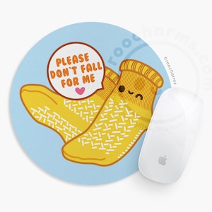 May include: A round mousepad with a light blue background featuring a cartoon illustration of two yellow socks with smiling faces. The socks have white grip patterns and are next to a white Apple mouse. Text reads "PLEASE DON'T FALL FOR ME".