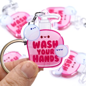 May include: Pink and clear acrylic keychain with a cartoon soap bottle and the text "Wash Your Hands". The keychain has a silver metal ring and a small clear bubble with a cartoon face.