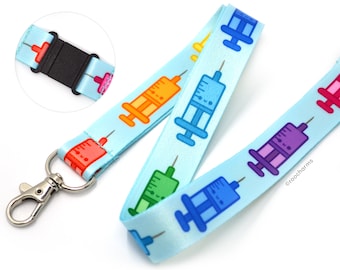 May include: A light blue lanyard featuring a repeating pattern of colorful cartoon syringes. The lanyard has a black plastic clasp and a silver-colored metal clasp. The syringes are in shades of red, orange, green, blue, and purple.