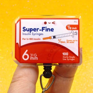 May include: A rectangular badge reel featuring a graphic of a Super-Fine insulin syringe box. The design includes the text Super-Fine Insulin Syringes, 1/2 Unit, 3/10mL, 30 units, 6mm, 31G, and 100 Sterile Single Use Syringes with a small face icon.