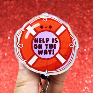 May include: A circular badge reel featuring a bright orange life preserver design with white accents and a small smiling face. The center of the life preserver is light purple and displays the text HELP IS ON THE WAY! in bold dark lettering.