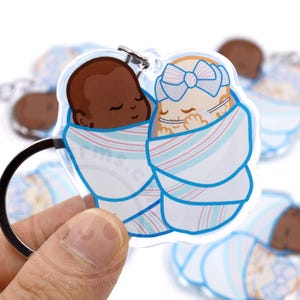 May include: A clear acrylic keychain featuring two cartoon babies wrapped in blue and pink striped blankets. One baby has dark skin, and the other has fair skin and a blue bow. The keychain has a silver ring.